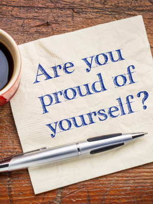 A handwritten note on a napkin reading “Are you proud of yourself?” placed beside a pen and a cup of coffee on a wooden table, symbolizing reflection and self-inquiry.
