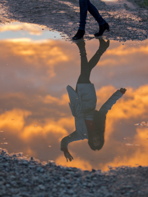 A reflection of a woman walking along a gravel path, seen upside down in a puddle reflecting golden clouds in the sky, symbolizing transition, uncertainty, and identity in between seasons.