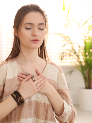 A woman with her eyes closed and hands resting over her chest, standing in soft natural light, symbolizing reflection, self-awareness, and an inward pause.