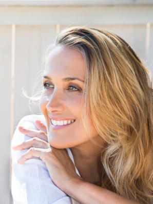 A woman with long blonde hair smiles softly while looking off into the distance, resting her hands near her face in natural light, conveying calm presence and contentment.
