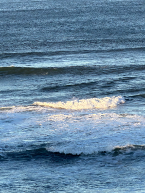 Soft ocean waves roll toward the shore along the Oregon Coast, with layered blue water and gentle white foam creating a calm, reflective coastal scene.